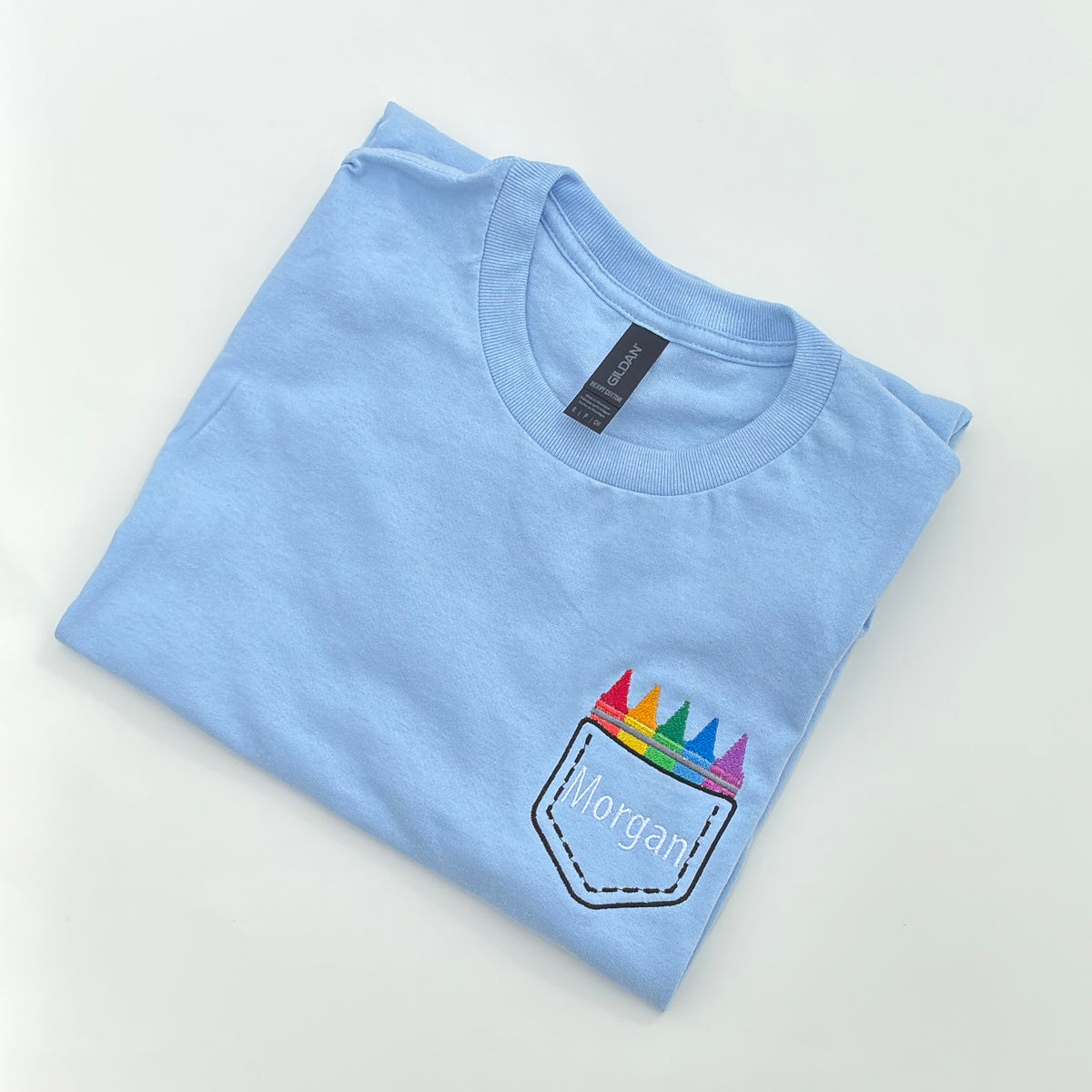 A light blue t-shirt with an embroidered design that resembles a faux crayon pocket on the chest area.