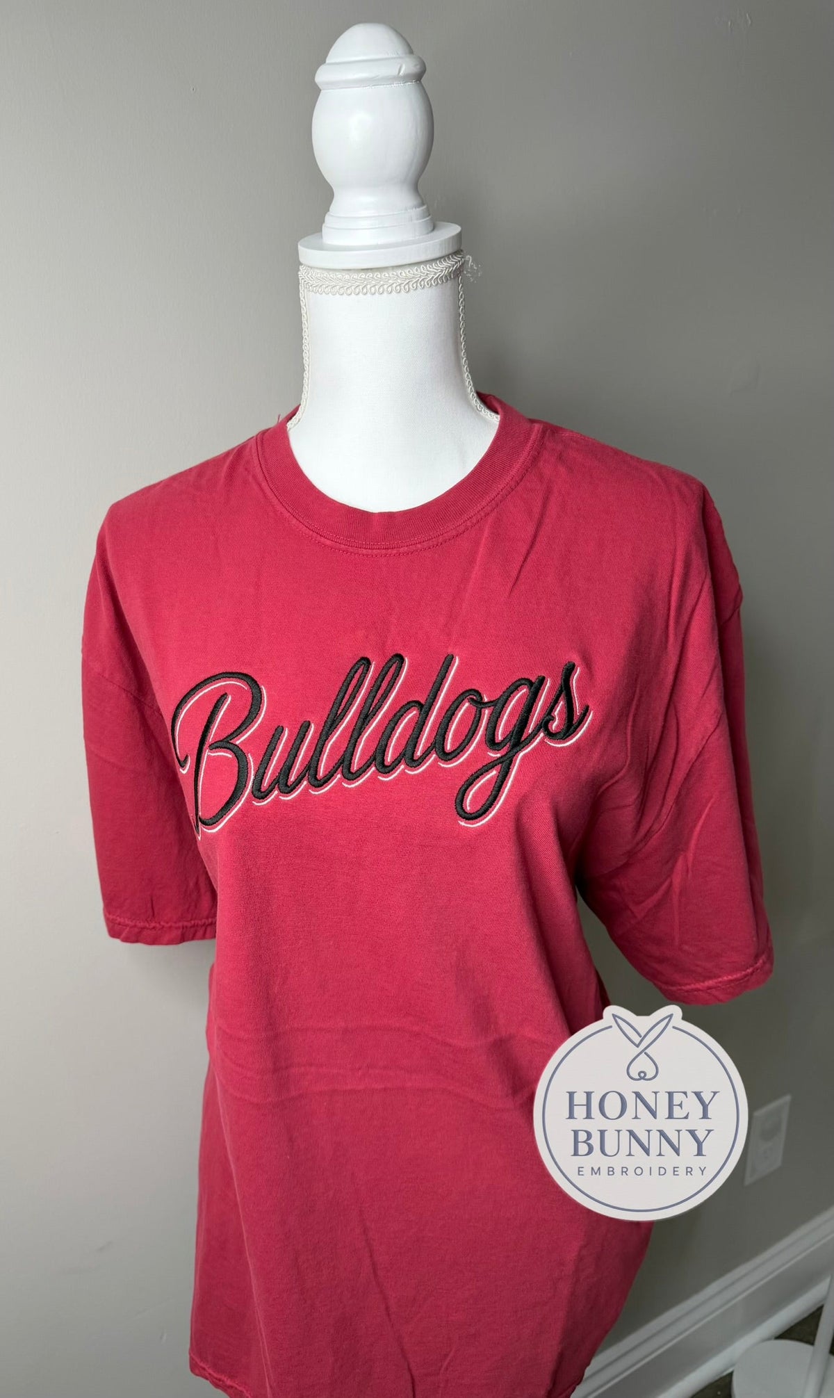 A pink short-sleeve t-shirt with 'Bulldogs' embroidered in black script with a white outline across the chest, displayed on a white mannequin torso. A 'Honey Bunny Embroidery' logo is visible in the bottom right corner.