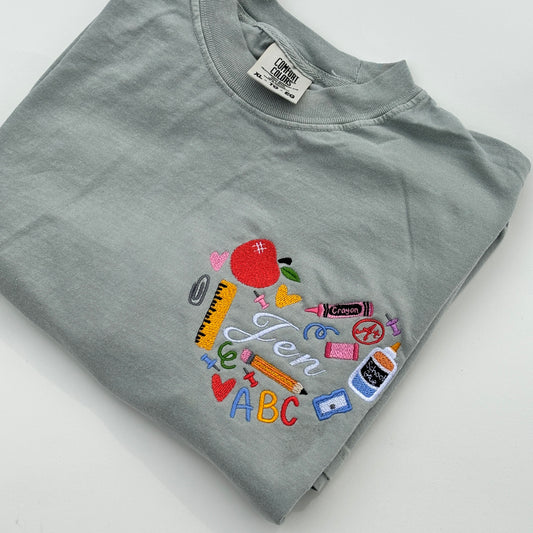 A green t-shirt with an embroidered design featuring various school supplies a