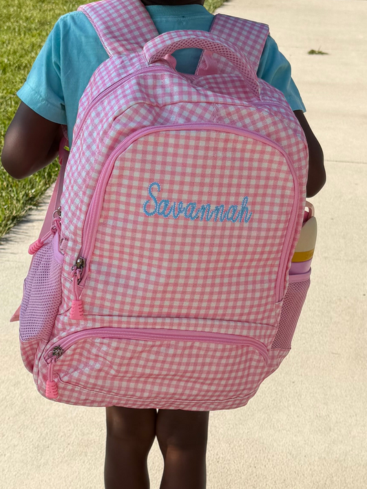 A child viewed from behind wearing a pink and white gingham backpack with the name 'Desiree' written in blue script on the front, standing on a concrete sidewalk in front of a brick building.