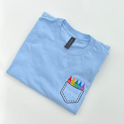 A light blue t-shirt with an embroidered design that resembles a faux crayon pocket on the chest area.