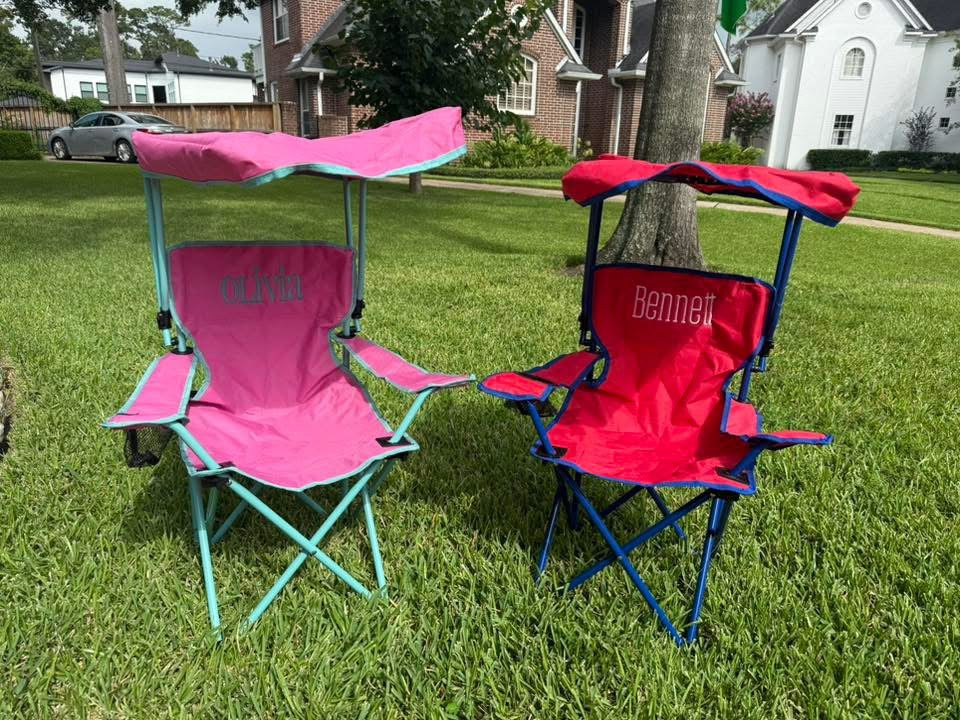 Two children's folding chairs with canopies, one pink and one red, on a grassy lawn.