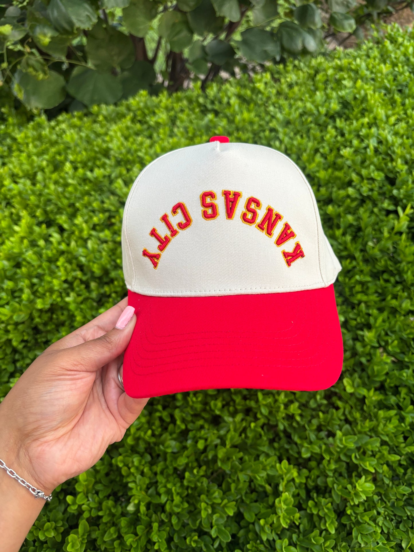 Red and white cap with 'Kansas City' text held by a hand against a green bush background