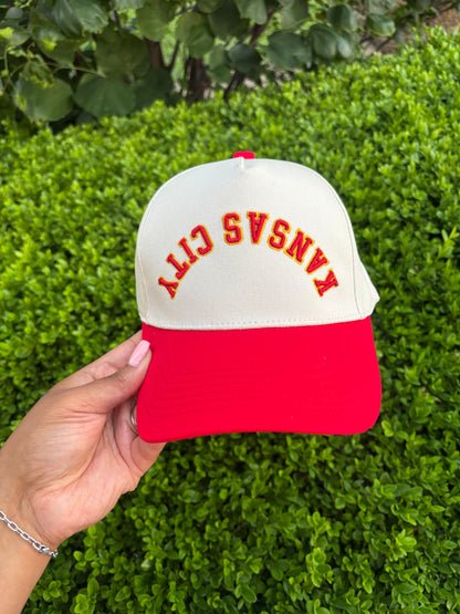 Red and white cap with 'Kansas City' text held by a hand against a green bush background