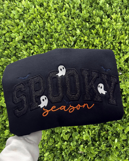A black sweatshirt featuring 'SPOOKY season' embroidered text with white ghosts and small blue bats, held by a hand against a background of green foliage.