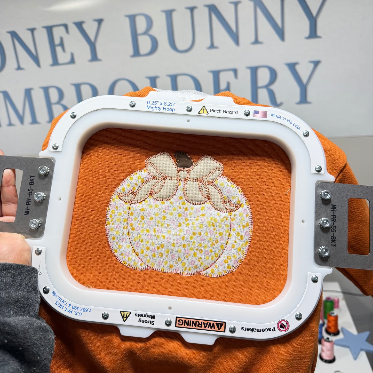 An embroidery machine hoop holding an orange fabric piece with a stitched pumpkin design featuring a cream and yellow floral pattern and a pink bow. A hand is holding the hoop, and the background shows a sign reading 'DONEY BUNNY EMBROIDERY'.