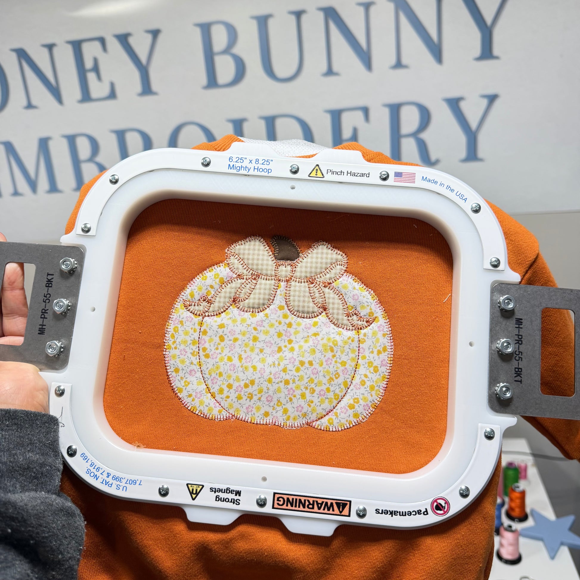An embroidery machine hoop holding an orange fabric piece with a stitched pumpkin design featuring a cream and yellow floral pattern and a pink bow. A hand is holding the hoop, and the background shows a sign reading 'DONEY BUNNY EMBROIDERY'.