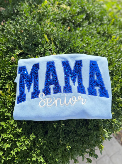 A light blue sweatshirt laid flat on green bushes, featuring the word 'MAMA' in large glittery blue letters and 'Senior' in white script across the front.