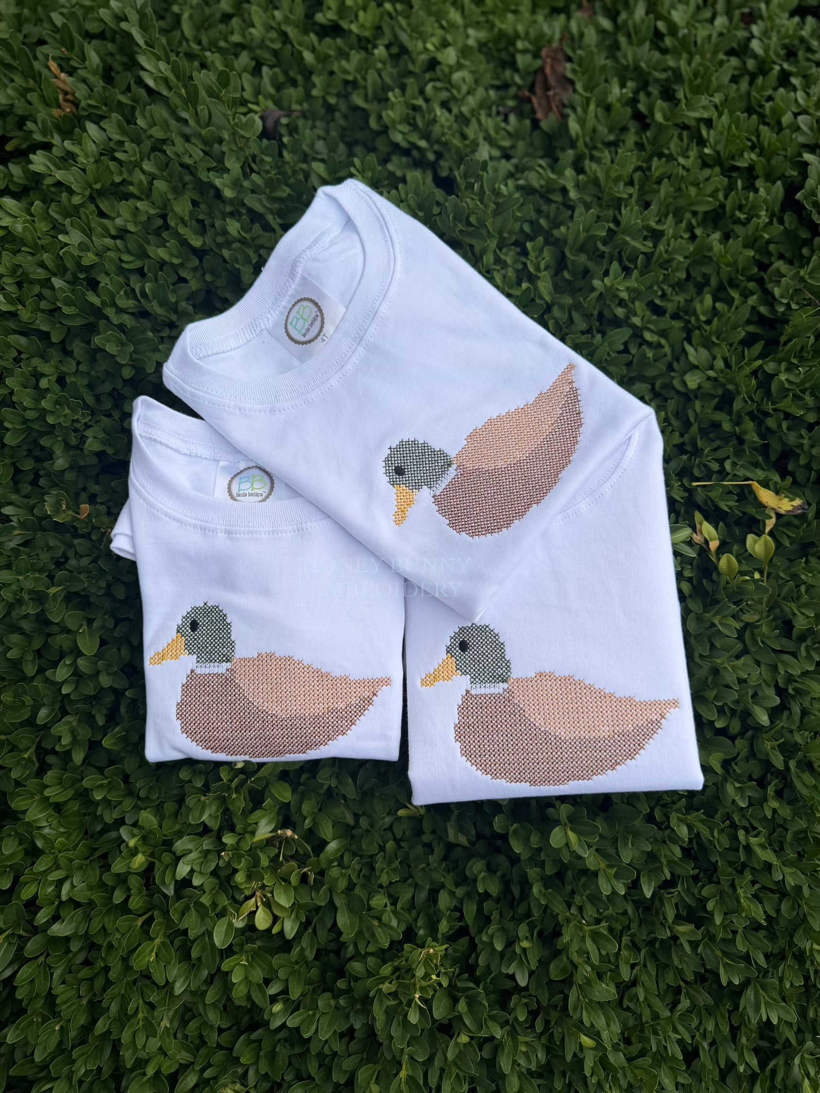 Three folded white long-sleeve shirts with embroidered ducks, arranged on a bed of green hedge leaves.