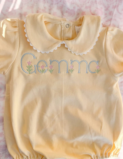 Yellow baby romper with 'Gemma' embroidered on it, laid flat on a pink surface.