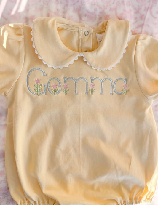 Yellow baby romper with 'Gemma' embroidered on it, laid flat on a pink surface.