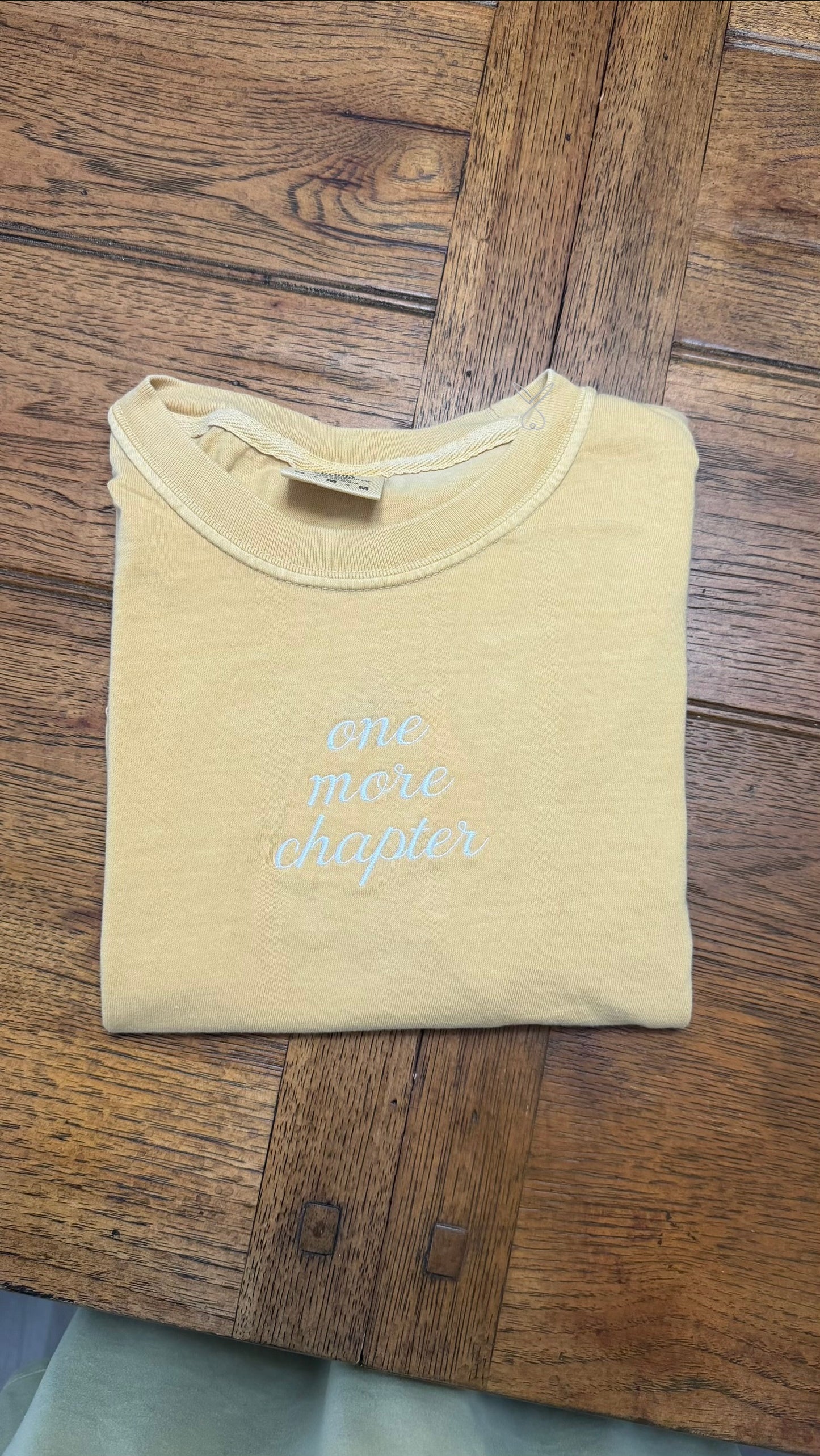 A folded mustard t-shirt with the phrase 'one more chapter' printed in light blue cursive text, laid flat on a dark wood floor.