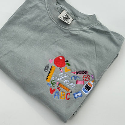 A green t-shirt with an embroidered design featuring various school supplies a