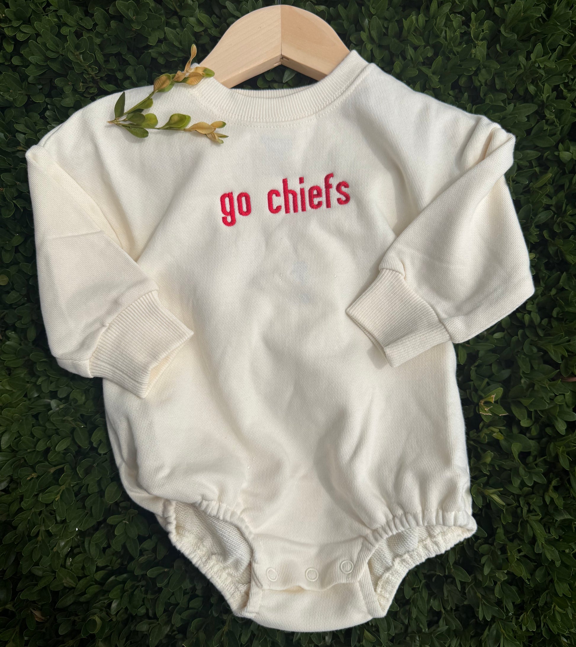 An off-white long-sleeve baby bodysuit with 'go chiefs' printed in red on the chest, displayed on a wooden hanger over a green hedge background.