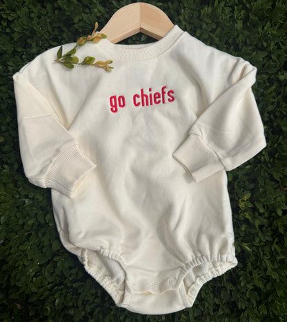 An off-white long-sleeve baby bodysuit with 'go chiefs' printed in red on the chest, displayed on a wooden hanger over a green hedge background.