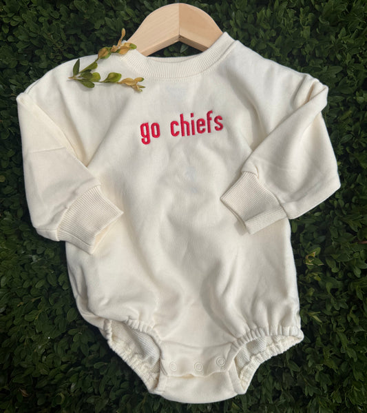 An off-white long-sleeve baby bodysuit with 'go chiefs' printed in red on the chest, displayed on a wooden hanger over a green hedge background.
