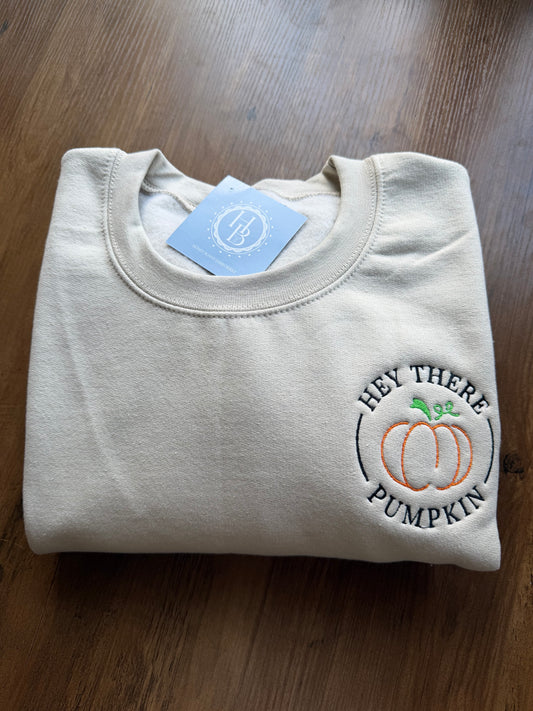 A folded cream-colored crewneck sweatshirt with an embroidered graphic on the left chest reading 'HEY THERE PUMPKIN' around an orange pumpkin illustration. A light blue brand tag is visible inside the collar, and the sweatshirt is laid flat on a dark wood surface.