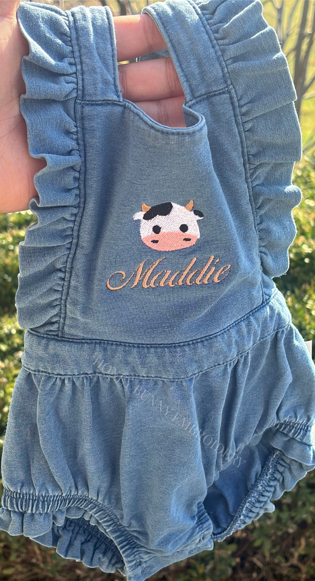 Blue denim romper with cow design and 'Maddie' text on a blurred natural background