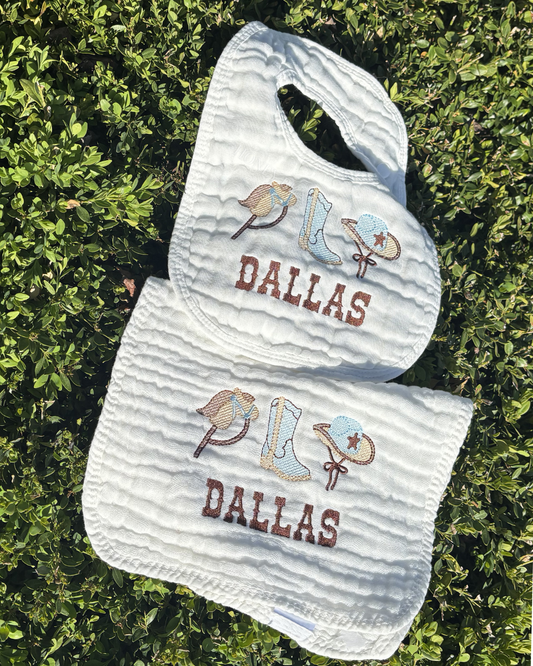 Baby bib and burp cloth set with 'Dallas' text and cowboy boot design on a green leafy background