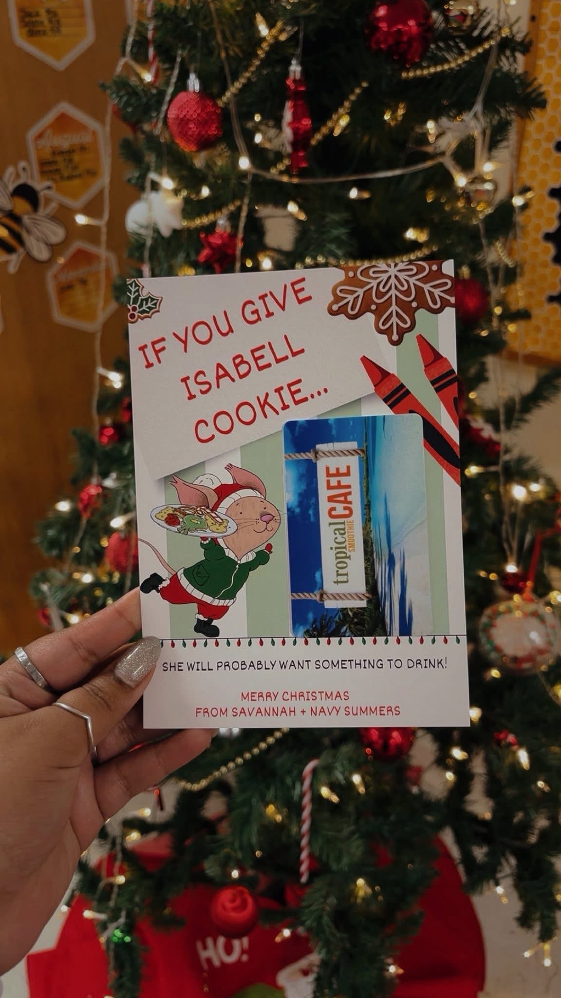 Hand holding a Christmas card with a cookie and drink reference in front of a decorated tree.