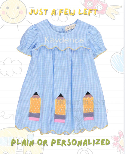 A light blue and white gingham baby dress with short puff sleeves, a ruffled neckline, and three applique pencil pockets on the front. The dress is shown against a white background with faint doodle graphics and text overlays reading 'JUST A FEW LEFT' and 'PLAIN OR PERSONALIZED'.