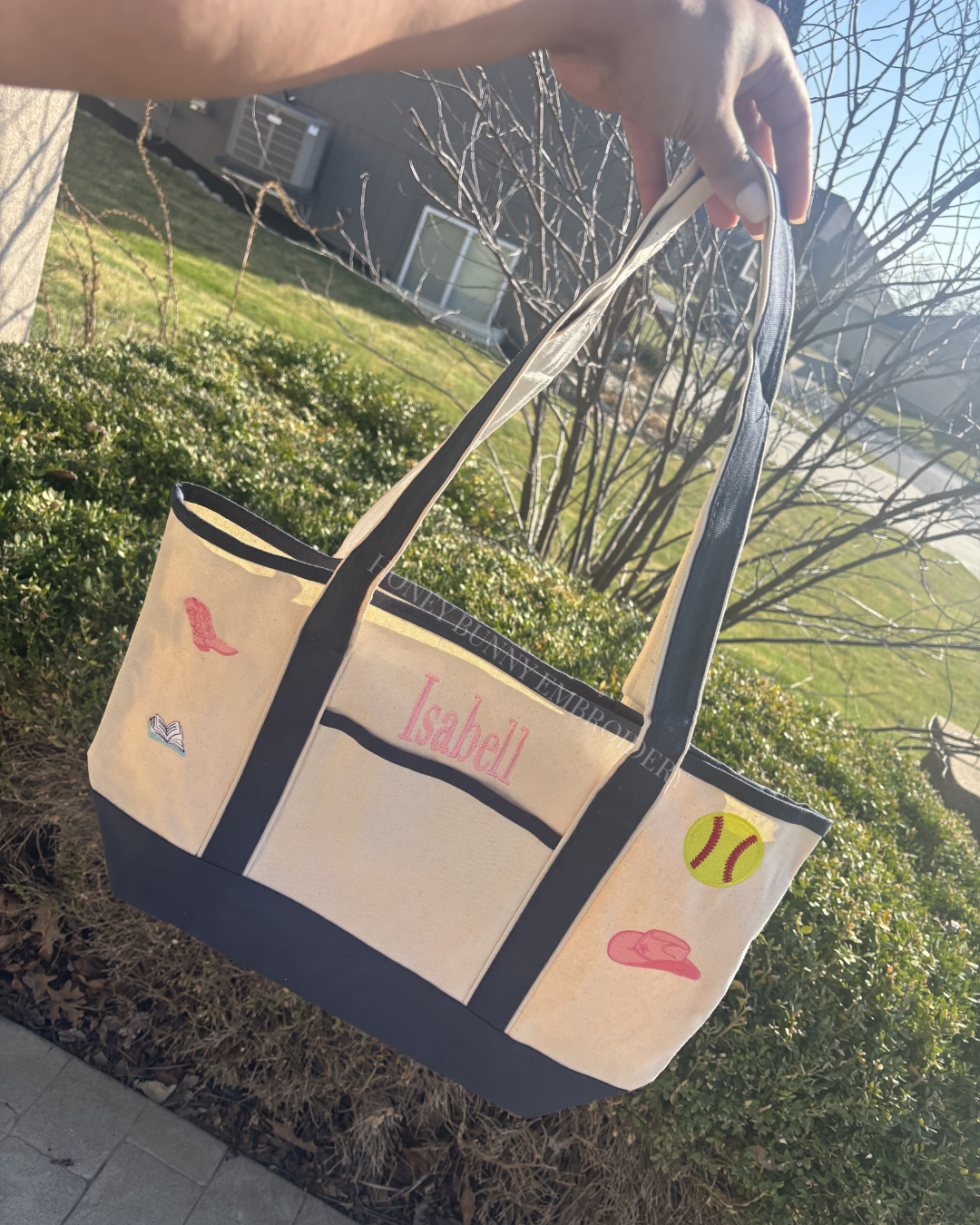 Beige tote bag with black trim, sports-themed designs, and 'Isabel' embroidered on it, held outdoors.
