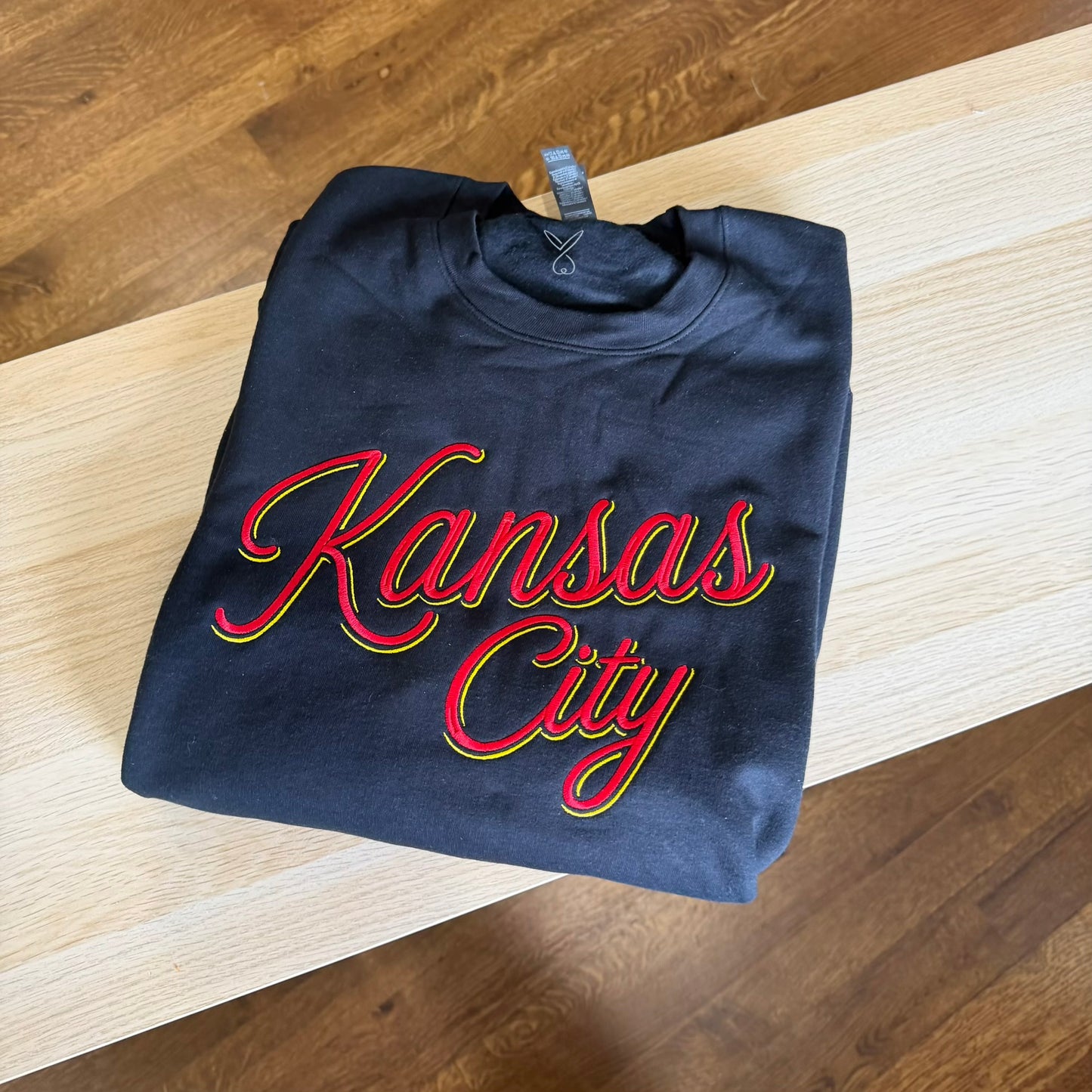 A folded black sweatshirt with 'Kansas City' written in red cursive script with a yellow and green outline, placed on a light wood table over a wooden floor.