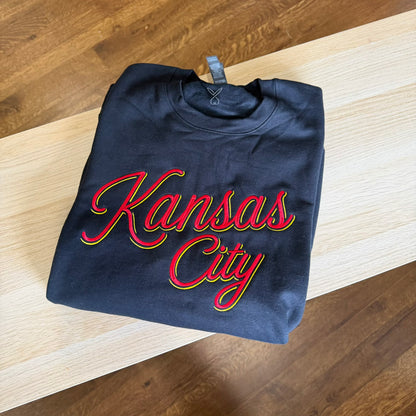 A folded black sweatshirt with 'Kansas City' written in red cursive script with a yellow and green outline, placed on a light wood table over a wooden floor.