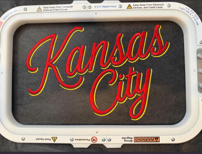 A rectangular vintage-style neon sign featuring the words 'Kansas City' in red cursive lettering with a yellow outline, set against a dark background and housed in a white metal frame with safety warning labels.