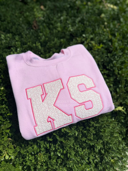 A folded light pink sweatshirt with large white varsity-style 'KS' letters outlined in pink, placed on green foliage.