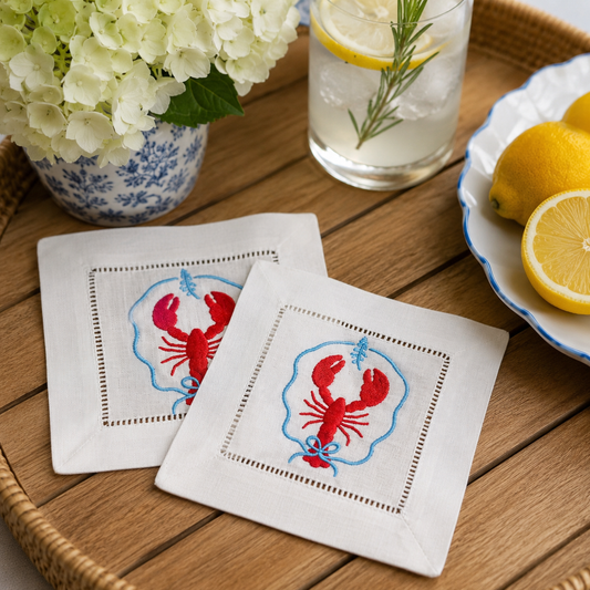 Two white napkins with red lobster design on a wooden table with a glass of lemonade and flowers.