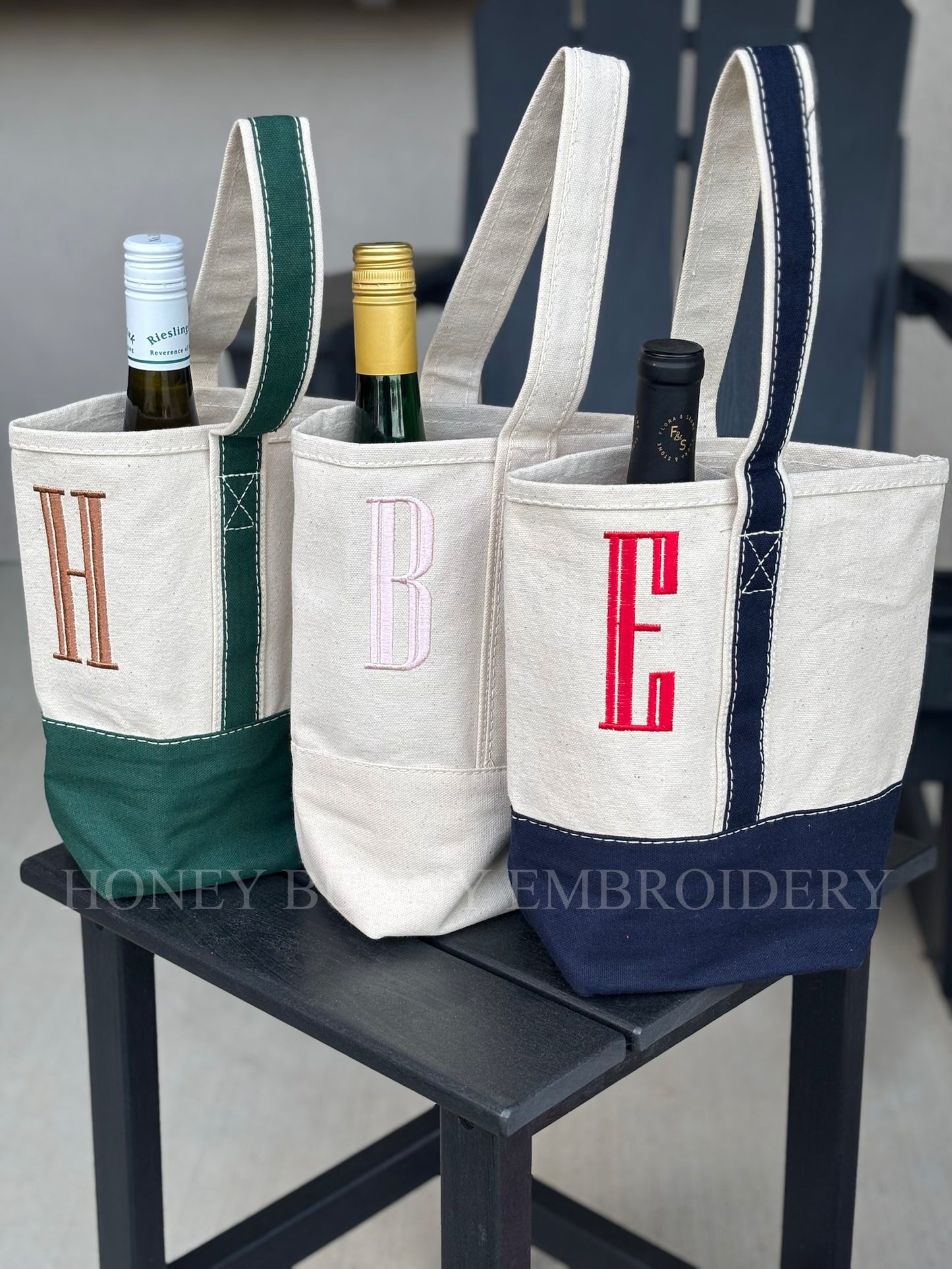 Three wine bottle bags with monogram letters on a chair, branded 'Honey Bee Embroidery'.