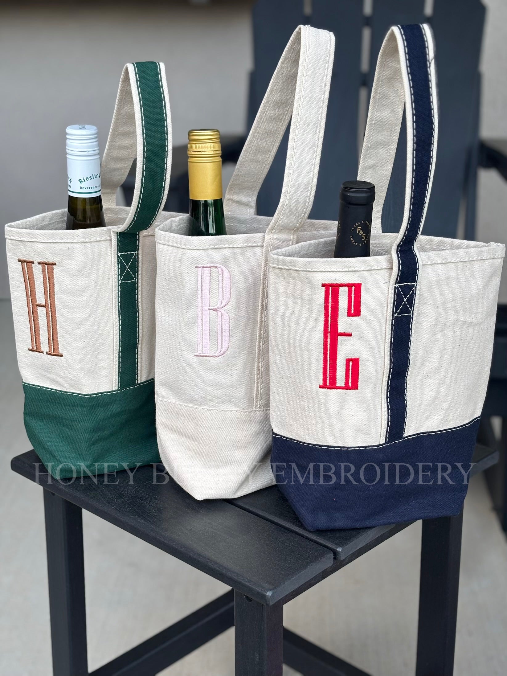 Three wine bottle bags with monogram letters on a chair, branded 'Honey Bee Embroidery'.
