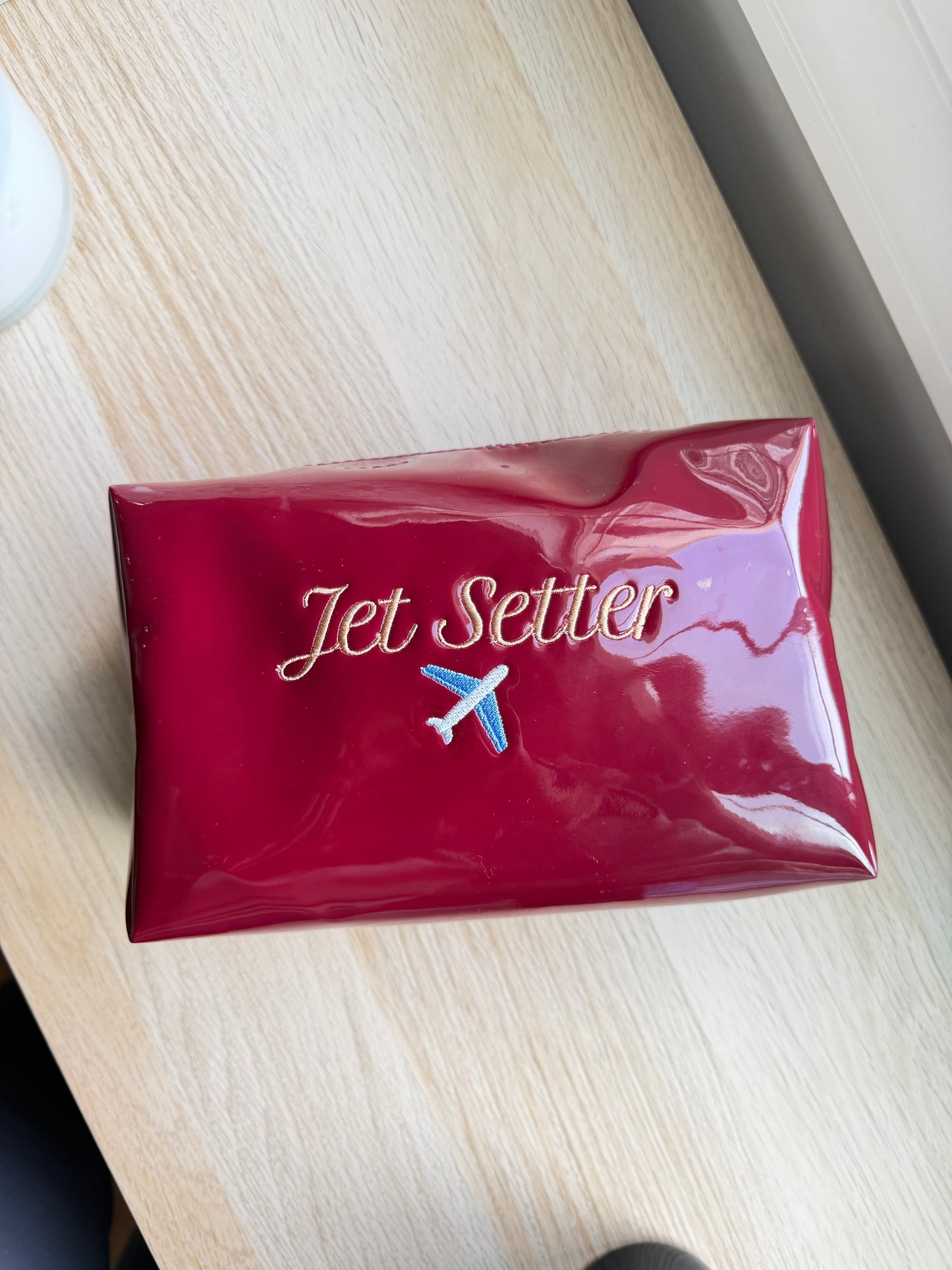 A glossy red rectangular pouch or case with the words 'Jet Setter' in gold script and a small airplane logo, placed on a light wood tabletop.
