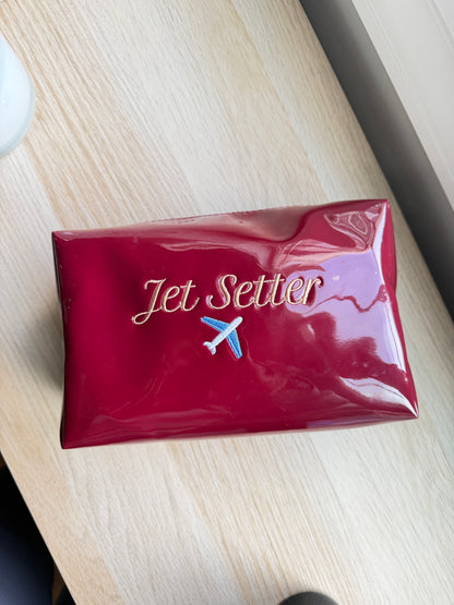 A glossy red rectangular pouch or case with the words 'Jet Setter' in gold script and a small airplane logo, placed on a light wood tabletop.