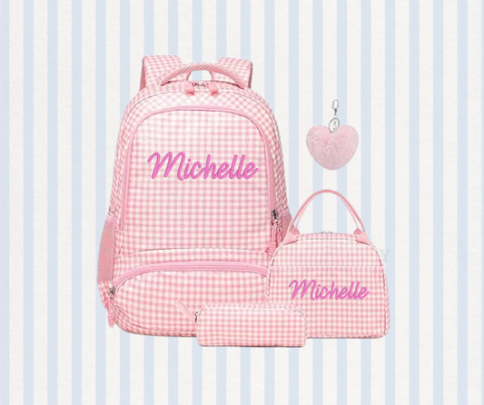A pink and white gingham backpack set with matching accessories, including a small pouch, a pencil case, and a heart-shaped keychain, all personalized with the name 'Michelle' in pink script. The set is displayed against a light blue and white striped background.