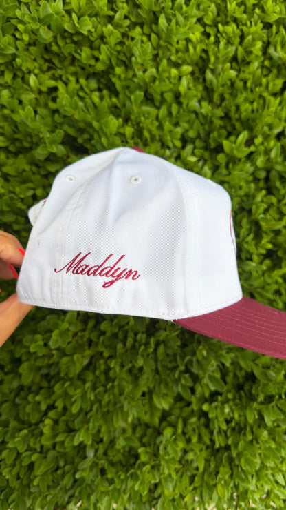White cap with red text held against a green hedge