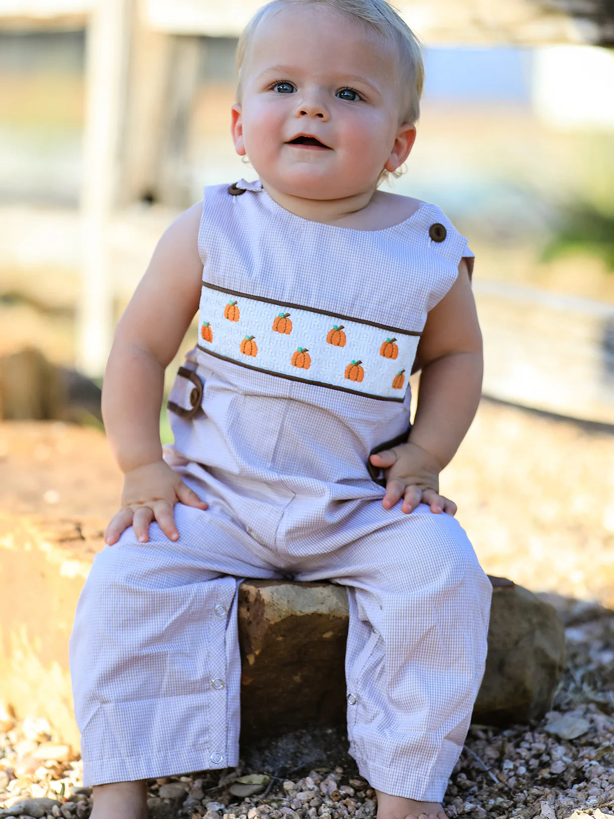 A baby sitting on a rock outdoors wearing a sleeveless light lavender overall romper with a horizontal white chest band decorated with orange pumpkins and dark brown trim.