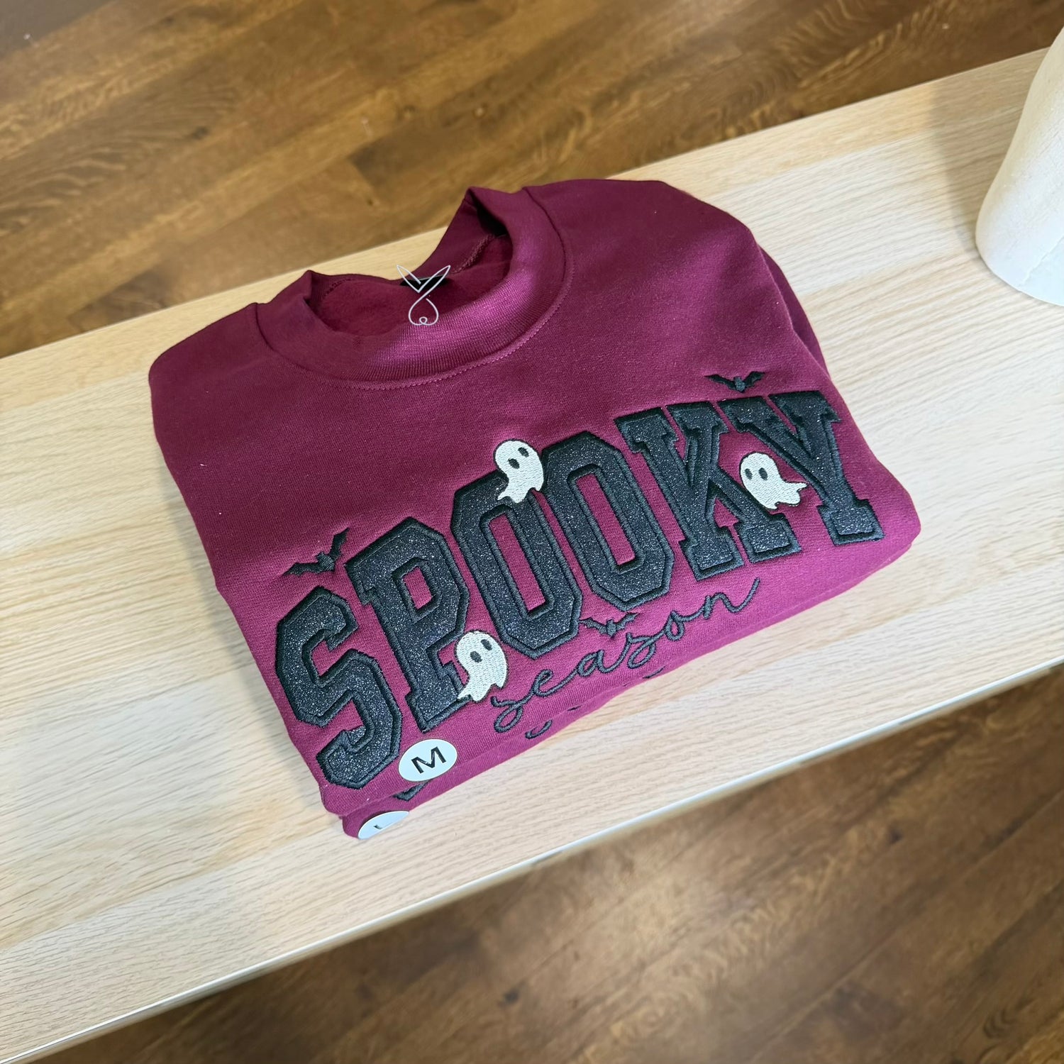 A folded maroon crewneck sweatshirt with 'SPOOKY season' embroidered in black and white across the front, placed on a light wood table over a dark wood floor.