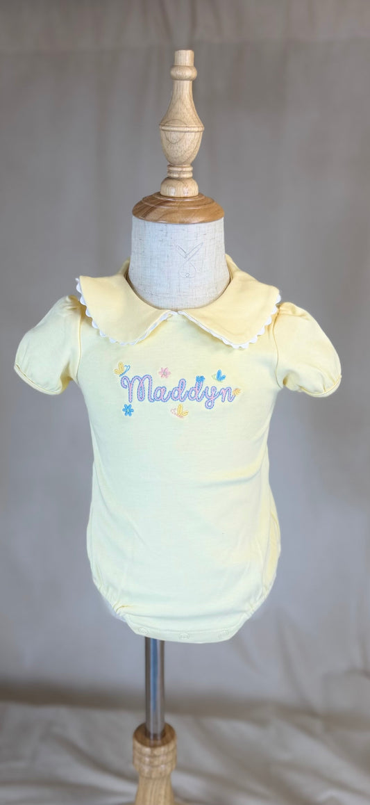 Yellow baby outfit on a mannequin with 'Maddox' embroidered on it against a plain background