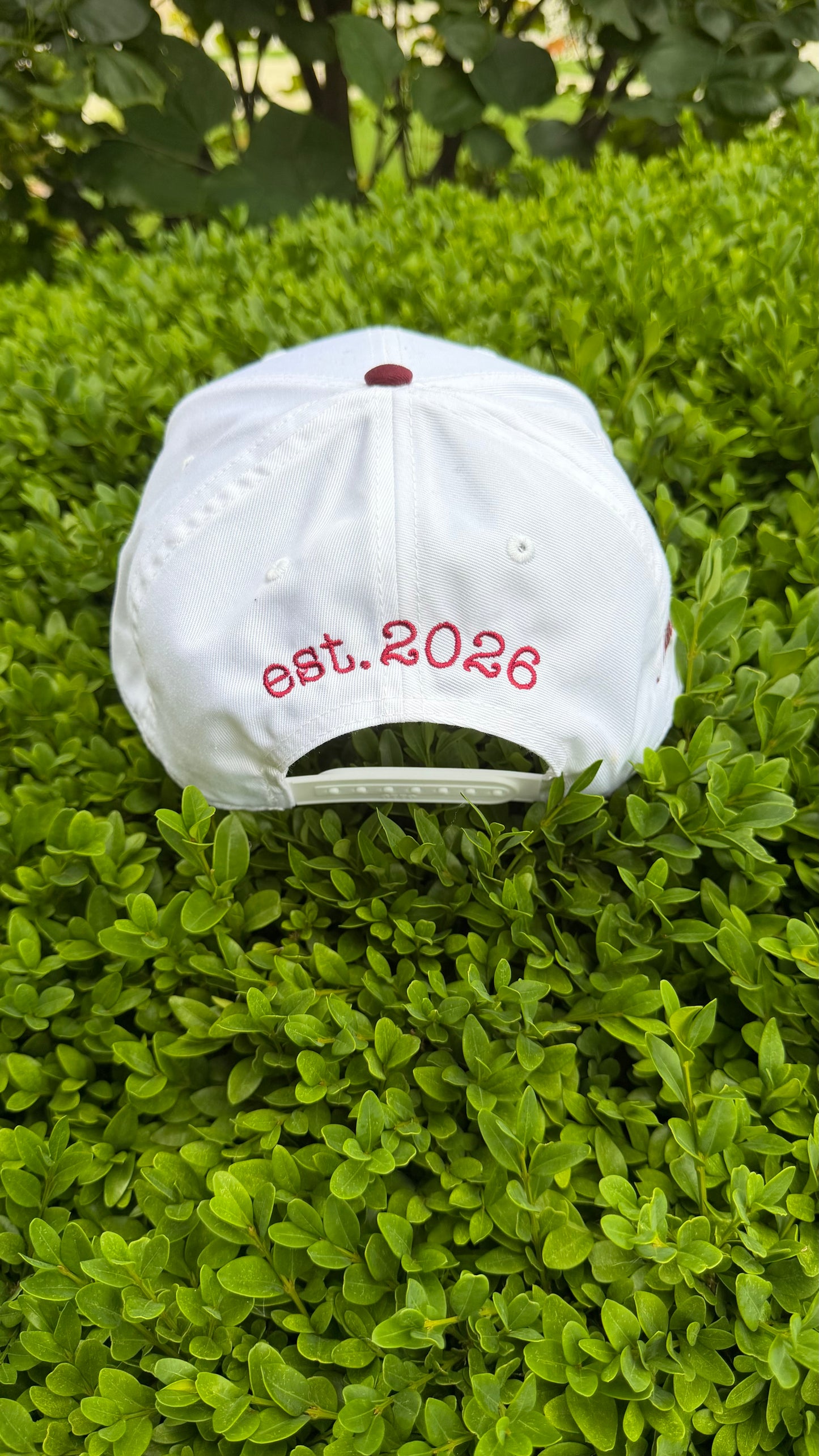 White cap with 'est. 2026' text on a green bush
