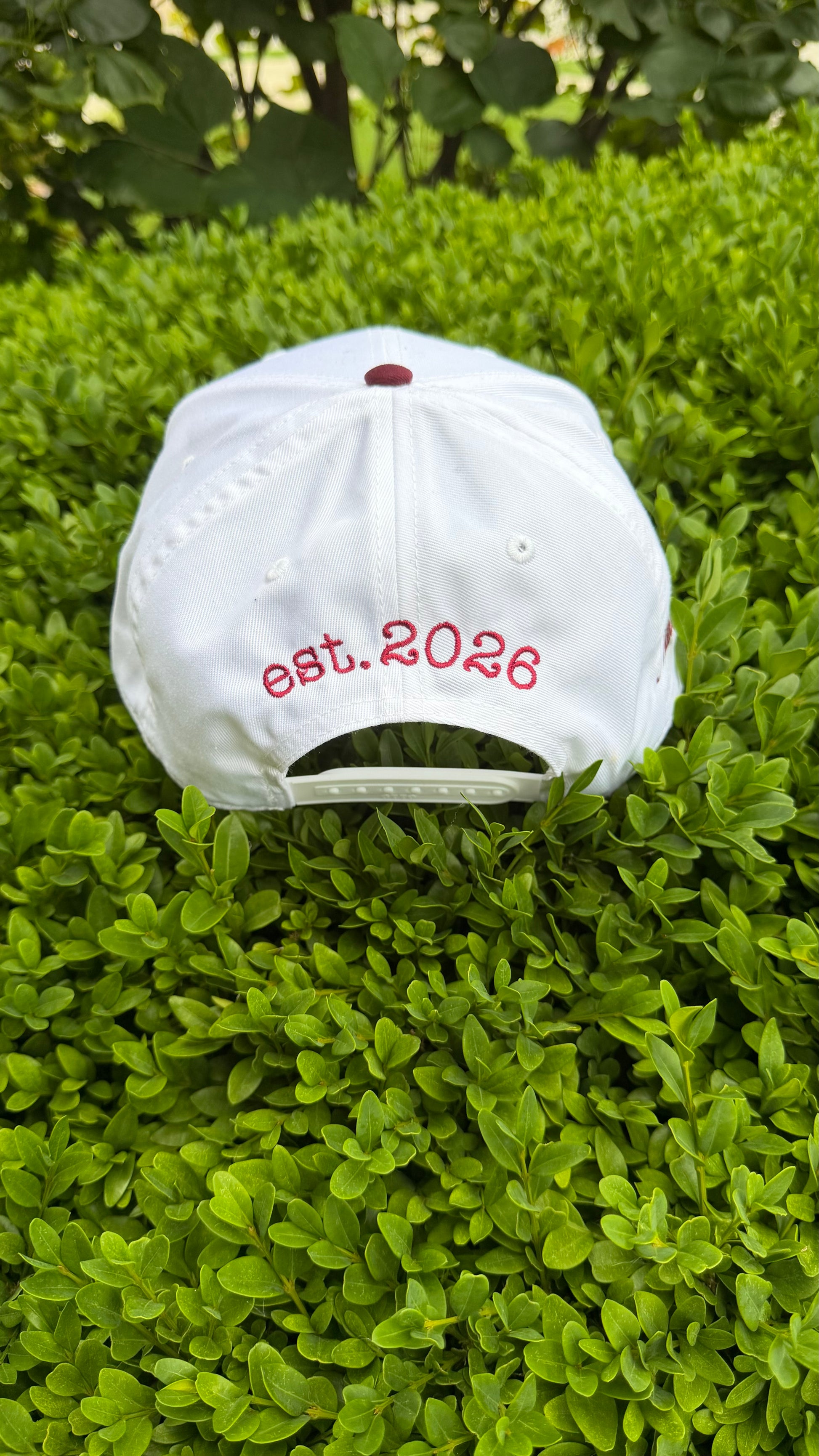 White cap with 'est. 2026' text on a green bush