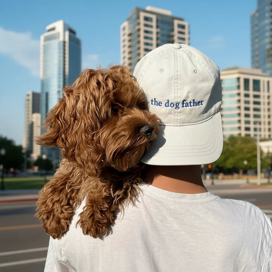Person wearing a cap with 'the dog father' text and holding a brown dog against a gray background