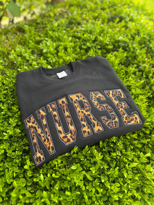 Black sweatshirt with 'NURSE' in leopard print letters on a green grass background