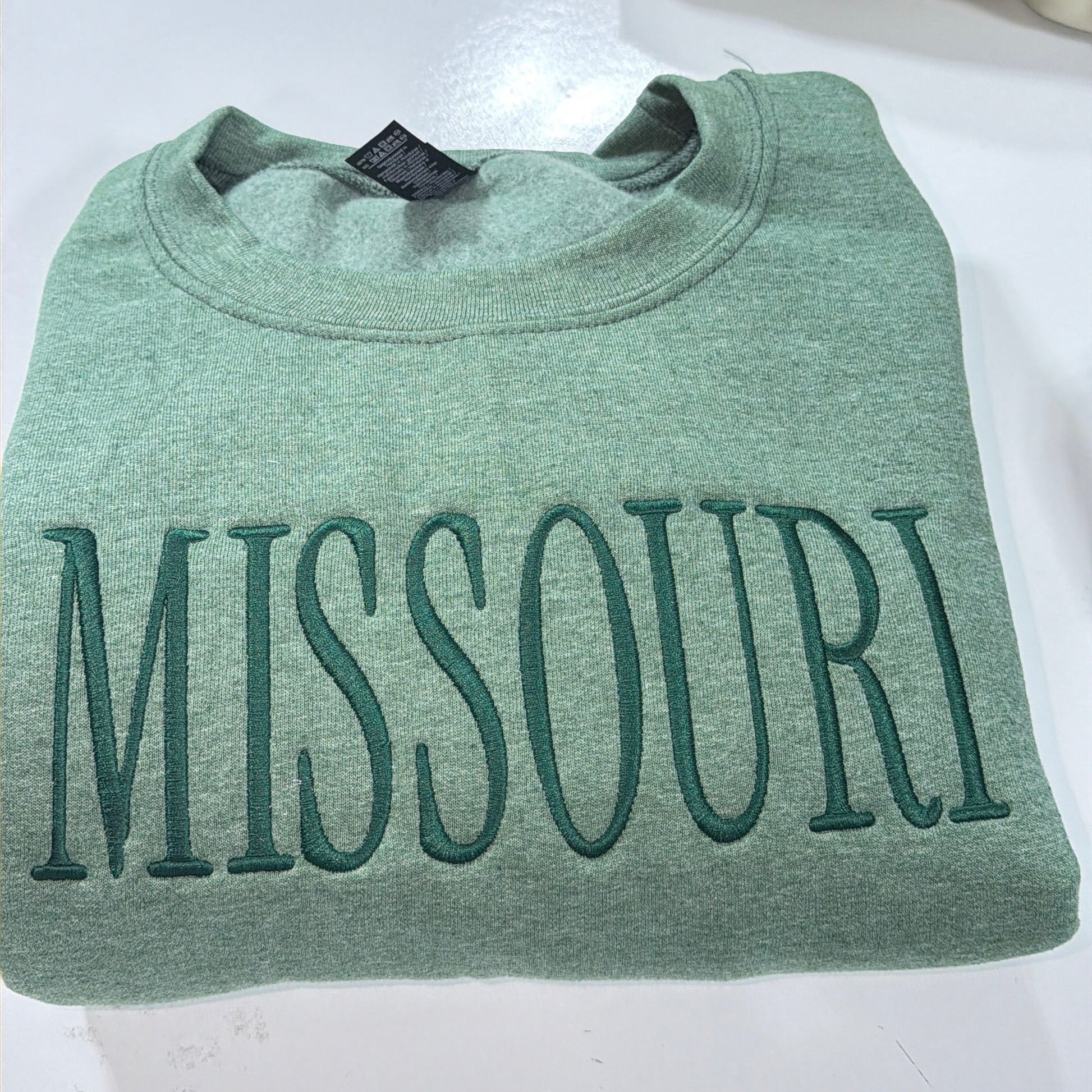Green sweatshirt with 'MISSOURI' printed on it, displayed with caps in the background.