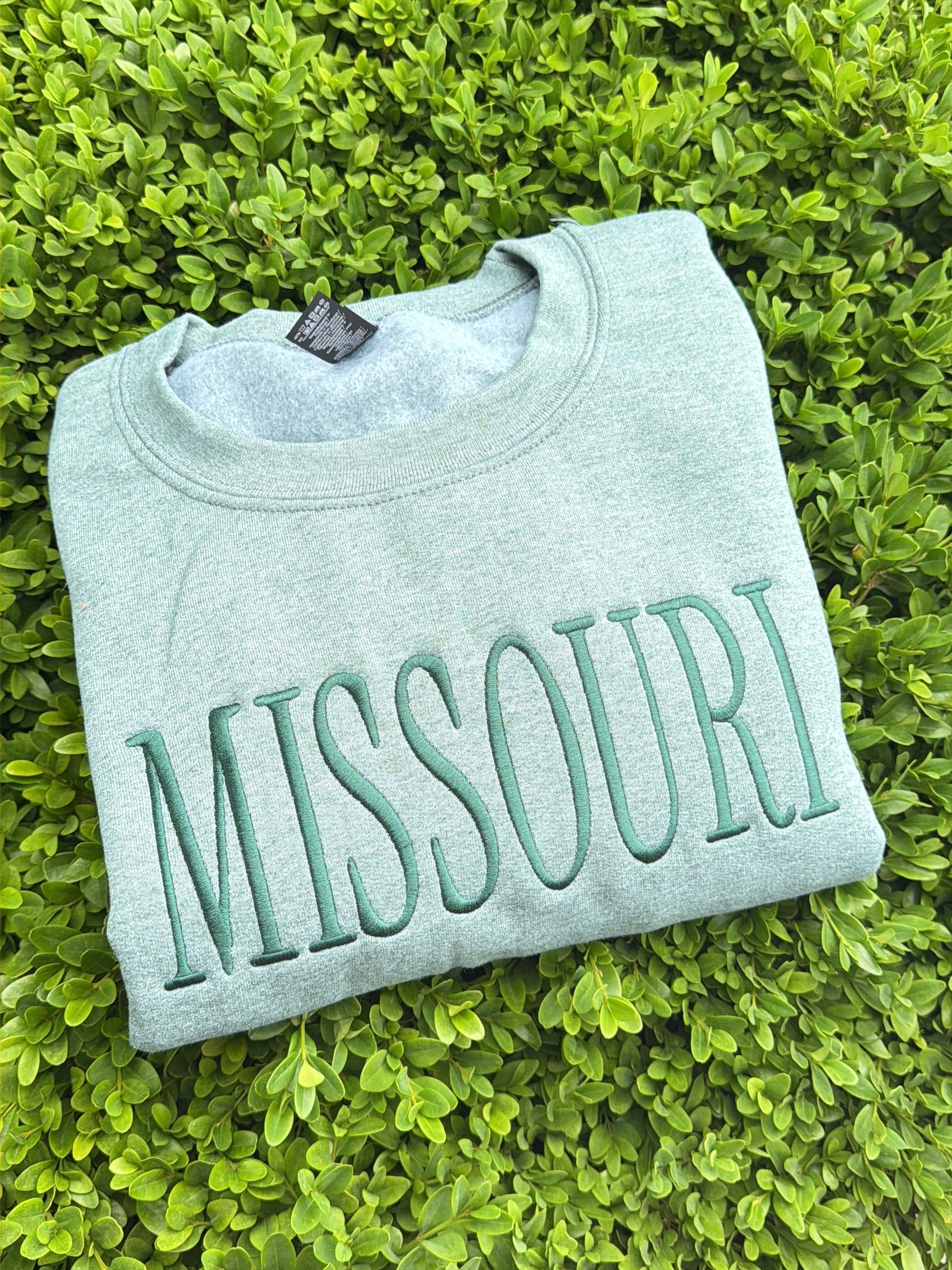 Folded heather green sweatshirt with 'MISSOURI' text on a green grass background midwestern pride