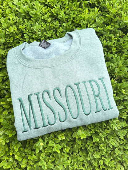Folded heather green sweatshirt with 'MISSOURI' text on a green grass background midwestern pride