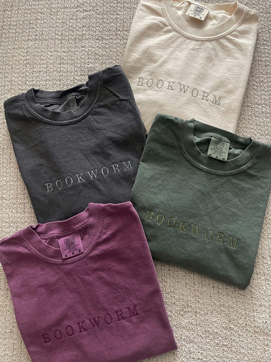 Four folded t-shirts in black, beige, green, and purple with 'BOOKWORM' text on a textured surface.