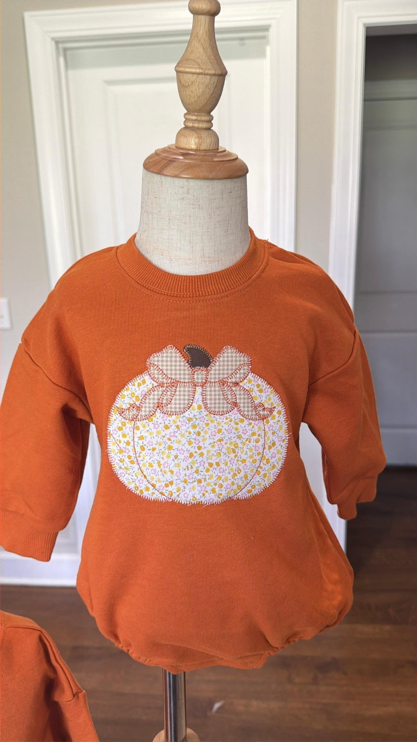 An orange crewneck sweatshirt displayed on a dress form mannequin, featuring a large applique pumpkin design on the front with a floral pattern and a bow.